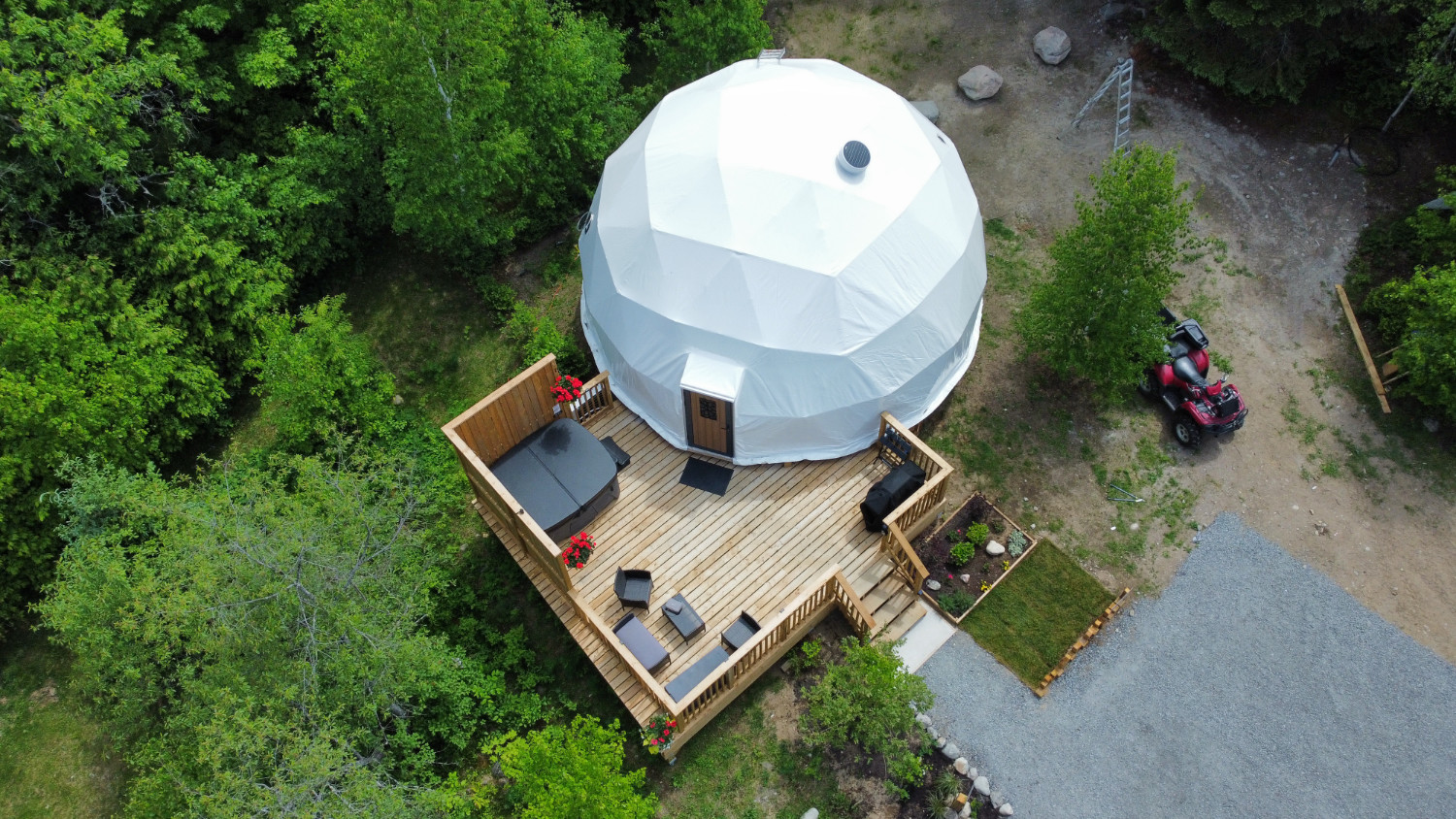 A white glamping dome site next to a wooden dock. With a red ATV beside it and green trees in the background. 